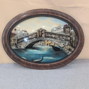 May include: Oval framed artwork depicting a bridge over a blue waterway, possibly Venice. The bridge is detailed with buildings on either side. The frame is a dark brown color.