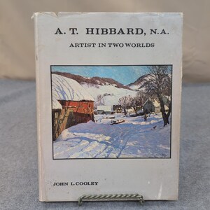 May include: A book cover depicting a winter scene with snow-covered fields and buildings. The title reads "A. T. Hibbard, N.A. Artist in Two Worlds" with the author's name, John L. Cooley, at the bottom. The cover shows signs of wear.