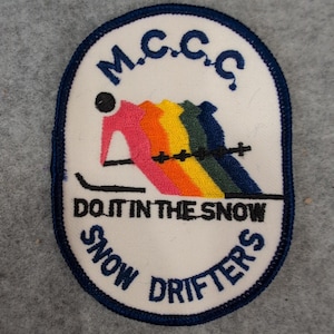 May include: Embroidered patch with a white background and a blue border. The patch features the text "M.C.C.C." at the top, and "DO IT IN THE SNOW" and "SNOW DRIFTERS" at the bottom. A skier in pink skis down a rainbow slope.