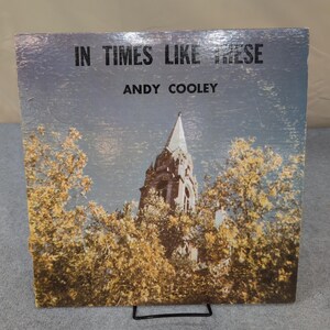May include: Vintage album cover with the text "IN TIMES LIKE THESE" and "ANDY COOLEY." The artwork shows a church steeple amidst autumnal trees with yellow and brown leaves, set against a blue sky.