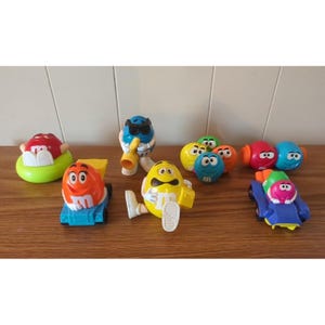 Puede incluir: Una colección de juguetes de personajes de M&M's coloridos. Los juguetes incluyen varios personajes de M&M's en diferentes poses, algunos con accesorios como gafas de sol e instrumentos musicales. Los juguetes están dispuestos sobre una superficie de madera.
