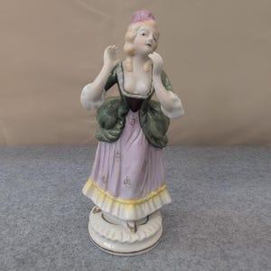 May include: A porcelain figurine of a woman in a historical dress. The figurine features a lavender dress with yellow trim, a green jacket, and a pink hat. The woman has blonde hair and is posed with her hands near her face. The base is white with gold accents.