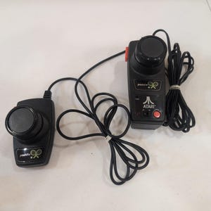 May include: Two black Atari paddle controllers with round knobs and red buttons. The controllers have the word "paddle" and a tennis racket graphic. The Atari logo is visible on one controller.
