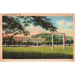 May include: Vintage postcard featuring Caserne de Nouméa NC. The image shows a large building with a red roof, surrounded by trees and a green field. The sky is a mix of blue and pink.