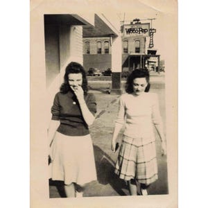Puede incluir: Fotografía vintage en blanco y negro de dos mujeres caminando por una acera. Una mujer lleva un suéter y una falda oscuros, la otra un suéter claro y una falda a cuadros. Un letrero de "Woco Pep" es visible en el fondo.
