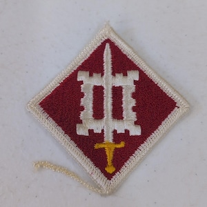 Op de afbeelding: Een ruitvormige patch met een rode achtergrond en een witte rand. Een wit kasteelachtig ontwerp is gecentreerd, met een zwaard dat er doorheen loopt. Het zwaard heeft een gele greep.