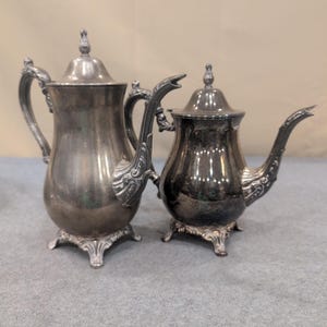 May include: Two antique silver teapots. The larger teapot has a curved handle and a decorative spout. The smaller teapot has a curved handle and a decorative spout. Both teapots have ornate bases and lids with finials. The teapots are displayed on a gray surface.