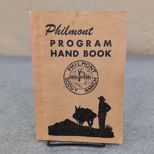 Puede incluir: Un libro antiguo de Philmont Program Hand Book con texto negro y un logotipo circular. La portada es de color beige con una silueta de un vaquero, un burro y un paisaje. El libro se muestra en un soporte de metal negro.