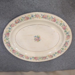 Pode incluir: Uma travessa de servir oval em cerâmica com uma cor base branco-sujo. A borda apresenta um padrão de guirlanda floral contínua com flores rosas, azuis e amarelas e folhas verdes. Um pequeno conjunto floral a condizer está centrado.