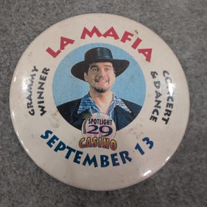 May include: A white button with the text "LA MAFIA" in red, and "Grammy Winner Concert & Dance" in black. The button features a man in a hat and the text "Spotlight 29 Casino September 13".