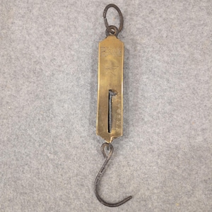 May include: A vintage brass spring scale with a hanging hook and a top ring. The scale has etched markings and the word "BARKE" above a star. The hook is dark metal, and the scale is rectangular.