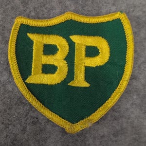 May include: A green shield-shaped patch with the yellow embroidered letters "BP". The patch has a yellow embroidered border. The background is a solid green color.