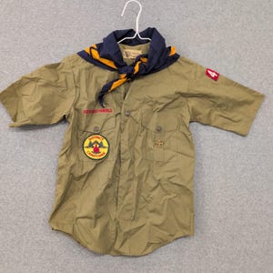May include: A khaki-colored Boy Scouts of America uniform shirt with short sleeves, two chest pockets, and a District Camporee patch. A blue and gold neckerchief is tied around the collar. The shirt has the number 4 on the right sleeve.