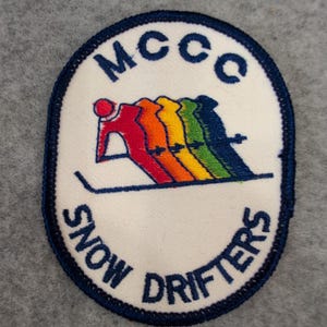 May include: An oval patch with a white background and a navy blue border. The patch features the text "MCCC" at the top and "SNOW DRIFTERS" at the bottom. In the center, a skier is depicted in a rainbow of colors.