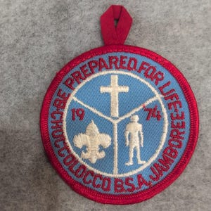 May include: A round patch with a red border and a red loop at the top. The patch has a blue center with white text that reads "BE PREPARED FOR LIFE JAMBOREE COLOCCO BSA". Inside the blue circle are a white cross, a fleur-de-lis, a figure, and the numbers "19" and "74".