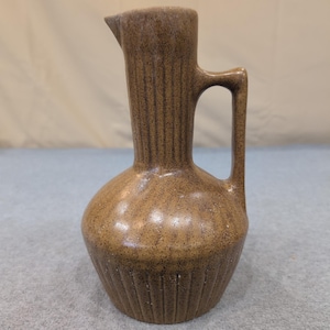 May include: A brown ceramic pitcher with a handle and a spout. The pitcher has a bulbous base and a tall, slender neck. Vertical lines decorate the neck and base. The pitcher is speckled with darker brown spots.