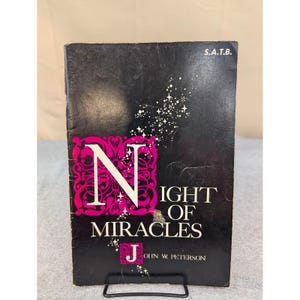 Pode incluir: Um livro vintage intitulado "Night of Miracles" de John W. Peterson. A capa é preta com uma grande letra "N" rosa e o título em branco. Desenhos de estrelas brancas e as letras "S.A.T.B." também são visíveis.