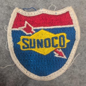 May include: Embroidered patch with a shield shape. The top section is red, the middle is blue, and the bottom is yellow with the word "SUNOCO". White stitching outlines the patch. Small red and white accents are on the patch.