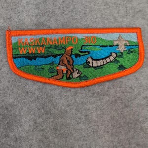 May include: Embroidered patch with an orange border. The patch features a colorful design with text that reads "KASKANAMPO 310 WWW". The image depicts a person, a river, a canoe, and a scout emblem.
