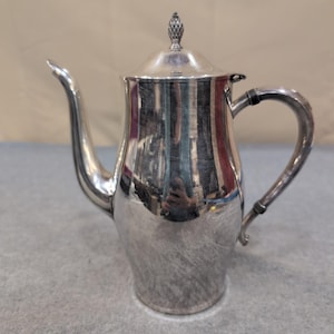 May include: A silver-colored coffee pot with a tall, slender body and a curved spout. The pot has a lid with a decorative finial and a curved handle. The surface reflects light, showing a polished finish. The pot is likely made of metal.