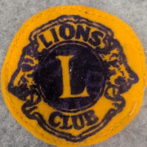 Może przedstawiać: Okrągła, żółta naszywka Lions Club z granatowym środkiem. Napis "LIONS CLUB" jest wygięty u góry i u dołu środka, z dużą żółtą literą "L" pośrodku. Naszywka jest wykonana z filcu.