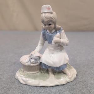 Statuetta vintage in porcellana raffigurante un'infermiera con un cucciolo in un cestino, nei colori blu e bianco. immagine 1