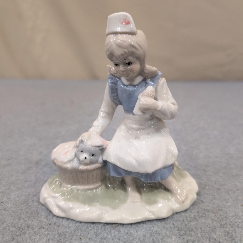 Statuetta vintage in porcellana raffigurante un'infermiera con un cucciolo in un cestino, nei colori blu e bianco. immagine 1