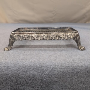May include: A silver-colored rectangular tray with ornate detailing. The tray has four curved legs and a decorative border with a repeating floral pattern. The tray is made of metal and has a reflective surface.