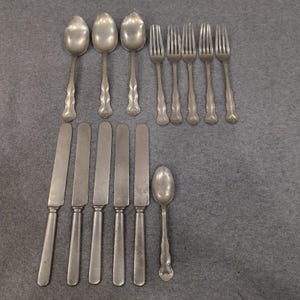 May include: A collection of antique silverware, including spoons, forks, and knives, arranged on a gray surface. The silverware has ornate handle designs and a tarnished silver finish, suggesting age and use. The arrangement includes various utensils for a formal dining setting.