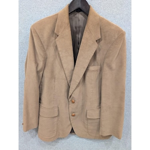 Puede incluir: Una blazer de pana color beige con solapa de muesca, dos bolsillos delanteros y dos botones marrones. La chaqueta está colgada de una percha negra sobre un fondo gris claro. La blazer tiene un diseño clásico y entallado.