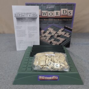 May include: A board game set of Upwords, a 3-dimensional word game. The set includes a green game board, letter tiles in a plastic bag, a rule sheet, and a box with the game title. The box reads "SCRABBLE UPWORDS".