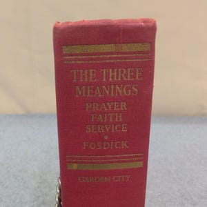 May include: A close-up of a red book spine with gold lettering. The title reads "THE THREE MEANINGS" with the words "PRAYER FAITH SERVICE" below. The author's name, FOSDICK, is also visible, along with "GARDEN CITY".