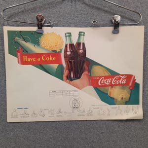 Könnte beinhalten: Vintage Coca-Cola-Werbung mit zwei Coca-Cola-Flaschen in einer Hand. Die Anzeige enthält den Text "Have a Coke" und "Drink Coca-Cola" auf roten Bannern. Ein Football und eine Uhr sind ebenfalls in der farbenfrohen Illustration zu sehen.
