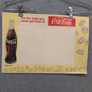 Könnte beinhalten: Vintage Coca-Cola Werbeplakat. Das Plakat zeigt eine Coca-Cola-Flasche, das Coca-Cola-Logo und den Text "For the taste you never get tired of." Das Plakat ist gelb und rot.