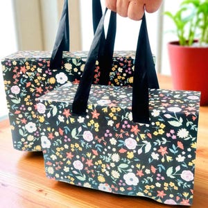 Op de afbeelding: Twee bloemenpatroon cadeau dozen met zwarte fluwelen handvatten. De dozen hebben een zwarte achtergrond met een kleurrijk bloemenpatroon. De bloemen zijn in tinten roze, geel en wit. De dozen worden vastgehouden, op een houten oppervlak.