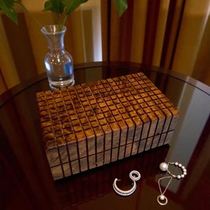 May include: A rectangular wooden box with a textured, grid-like pattern on the lid. A clear glass vase with a plant is in the background. Jewelry, including a pearl necklace, bracelet, and a diamond earring, is on the table.