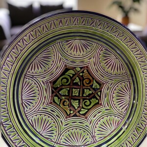 May include: A decorative ceramic plate with a detailed geometric design. The plate features a central star-shaped pattern in black, brown, and green, surrounded by concentric circles of green and purple fan-like motifs. The rim is dark blue.