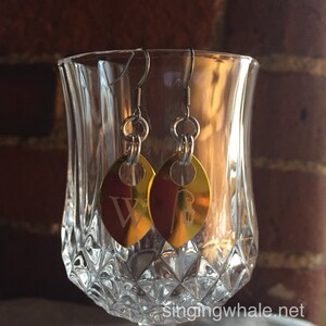 May include: A pair of gold-colored earrings with a leaf-shaped design. The earrings have a silver hook and a small letter 'W' engraved on one leaf and a small design on the other.