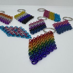May include: A collection of handmade keychains featuring colorful chainmail designs. The keychains display various rainbow and gradient color schemes, including pink, blue, purple, and yellow. Each keychain has a silver-colored keyring attached.