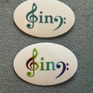 May include: Two white oval stickers with the text "sing:" and a treble clef symbol. The top sticker has a teal treble clef, while the bottom sticker has a rainbow-colored treble clef.