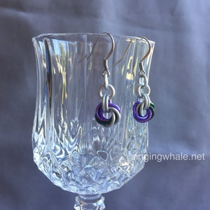 May include: A pair of silver earrings with a purple, green, and white woven design. The earrings are hanging from a glass goblet.