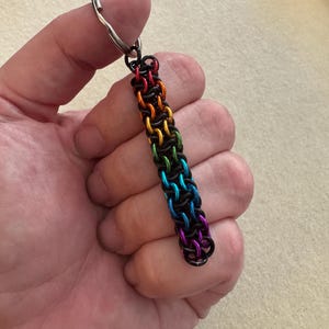 Op de afbeelding: Een sleutelhanger met een rechthoekige strook maliënkolder, opgebouwd uit in elkaar grijpende zwarte metalen ringen en levendige regenboogkleurige ringen. Het regenboogpatroon verloopt van rood bovenaan naar oranje, geel, groen, blauw en paars.