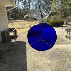 May include: A stained glass peace sign in a vibrant blue color, suspended by a suction cup. The peace symbol is divided into three sections by black lines. The sun shines through the glass, creating a bright and colorful display.
