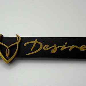 Può includere: Un portachiavi rettangolare nero con la scritta "Desire" in oro. Un emblema dorato è sul lato sinistro. Il portachiavi ha un portachiavi argentato.