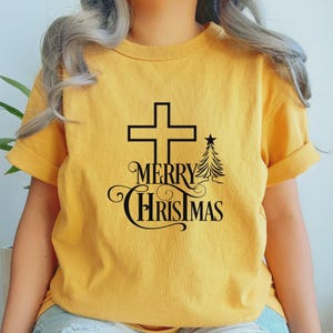 May include: Mustard yellow t-shirt with a black graphic. The graphic includes a cross, a Christmas tree, and the words "MERRY CHRISTMAS" in a decorative font. The shirt has rolled-up sleeves.
