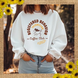 Op de afbeelding: Witte sweatshirt met de tekst "REGISTERED TECHNICIAN BEHAVIOR Coffee Club 1985". Het ontwerp bevat een rennende koffiekop-afbeelding. De tekst "MAY YOUR COFFEE BE STRONGER THAN YOUR MONDAY" staat ook op de sweatshirt.