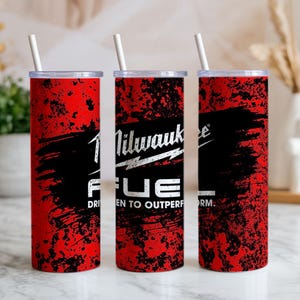 May include: Three cylindrical tumblers with a red and black distressed texture pattern. Each features the white Milwaukee Fuel logo and the text Driven to Outperform. The tumblers have clear lids and white straws, standing upright in a row.