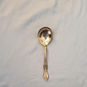 May include: A gold-colored spoon with an oval bowl and a decorative handle. The spoon has an ornate floral design on the handle and bowl. The spoon is likely made of metal and is a piece of silverware.