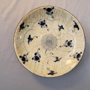 May include: Round ceramic plate with a cream-colored background and a cracked glaze effect. The plate is decorated with blue floral motifs and a central spiral design. The rim is dark blue.