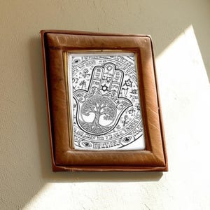 Puede incluir: Una impresi&oacute;n art&iacute;stica en blanco y negro enmarcada de una mano de Hamsa. La obra de arte presenta una ilustraci&oacute;n detallada de una mano con un ojo en la palma, rodeada de s&iacute;mbolos y un dise&ntilde;o de &aacute;rbol de la vida. El marco es de color marr&oacute;n c&aacute;lido.